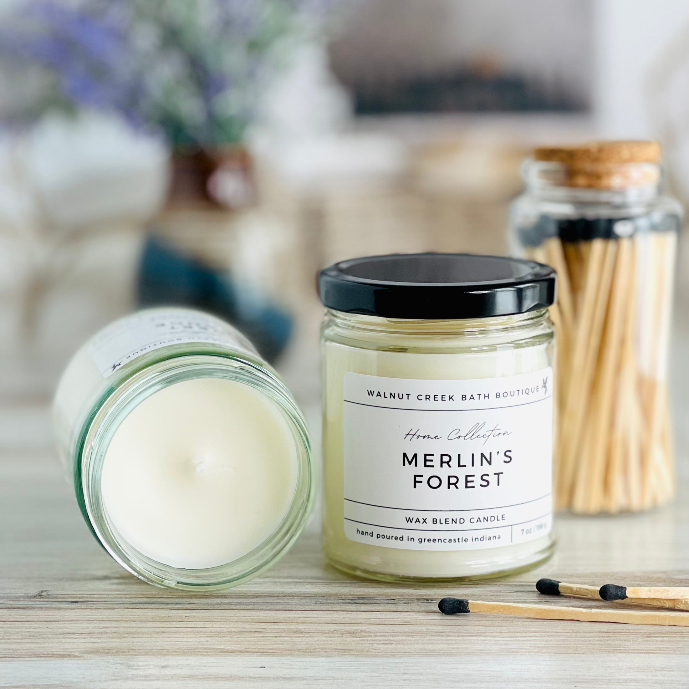 merlins forest candle with lid to the right of another candle with the top off to show the creamy wax inside. there are a few matches scattered in front and a pretty glass jar of matches in the background