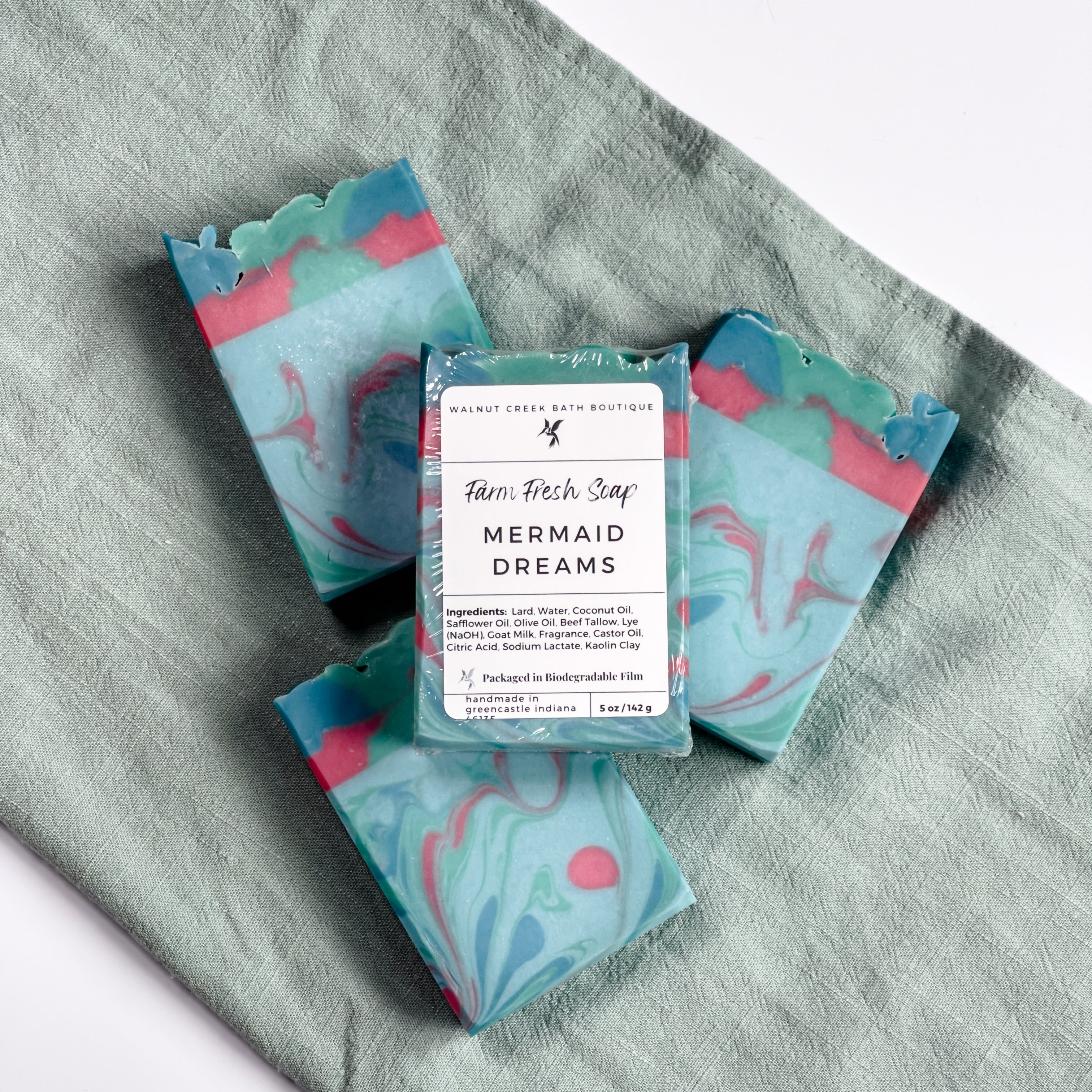 3 mermaid dreams soaps are laying flat on a green cloth with another on top of them showing the label.