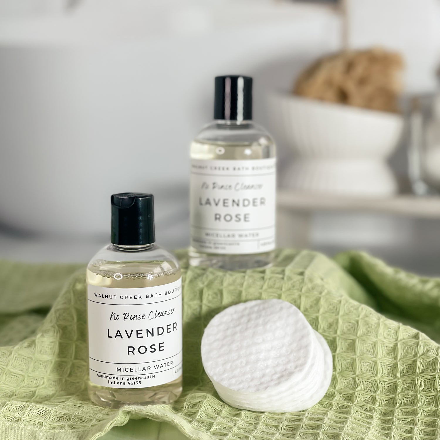 2 micellar waters are shown on a green textured cloth covered riser along with some makeup remover pads. The background is a spa like bathroom