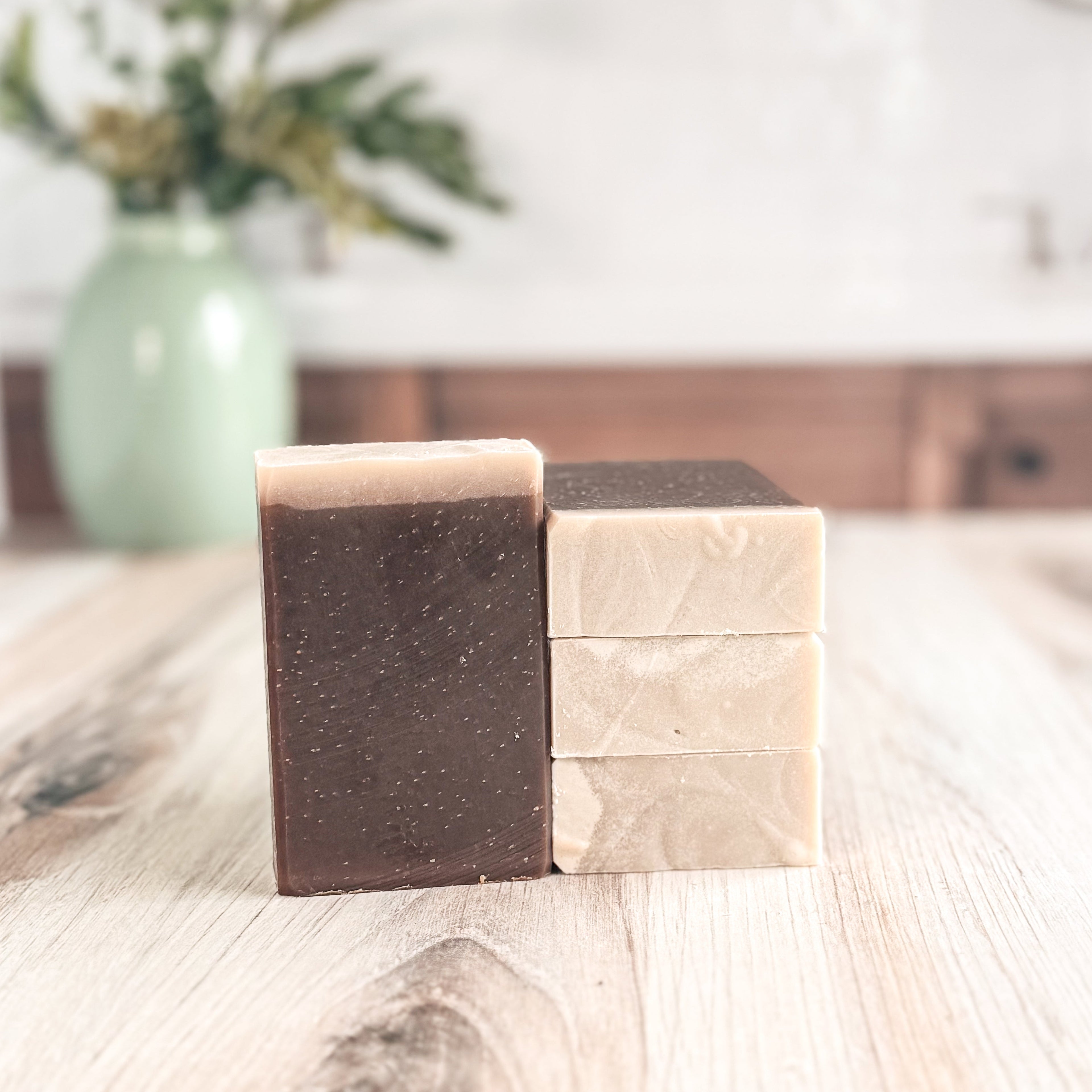 Two bars of soap, one brown and one beige, on a wooden surface with a blurred background.