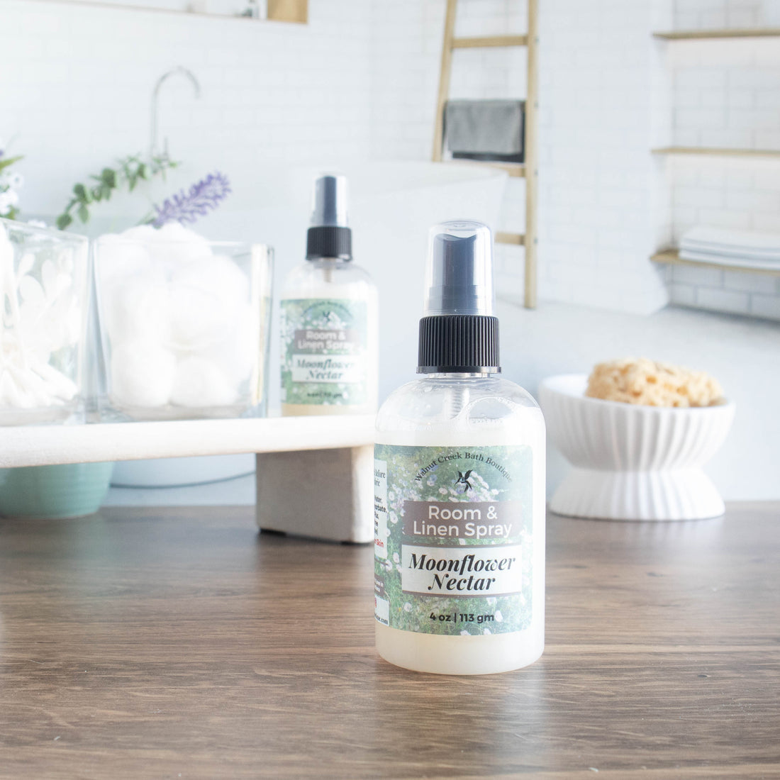 moonflower nectar room spray is front and center. it is in a squat round bottle with black sprayer. in the background is a tray with some cotton balls and q-tips in jars along with another room spray. in the background is an image of a spa bathroom and a dish with a loofah