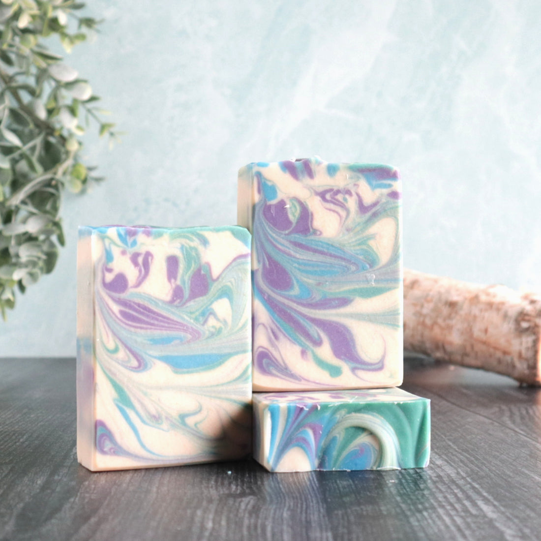 3 bars of perfectly manly soaps are pictured. The soaps have a white base with blue, green and purple swirls. There is some greenery peaking out in the left and some small logs in the back right. The background is a pretty blue-green color.
