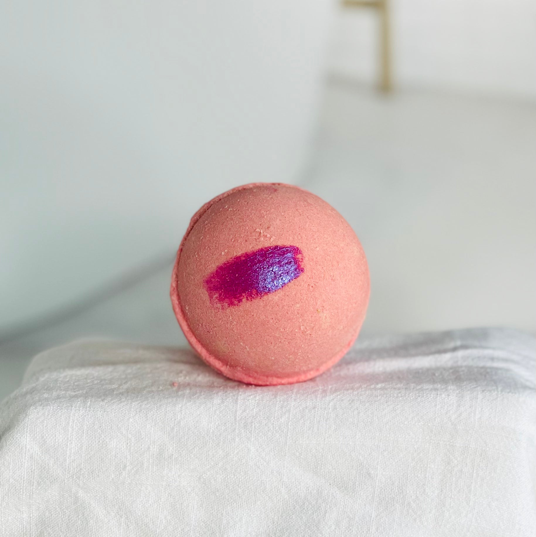 close up view of pink sands bath bomb. the bath bomb is pink with a much darker pink slash. this is sitting center stage on a white cloth over a riser