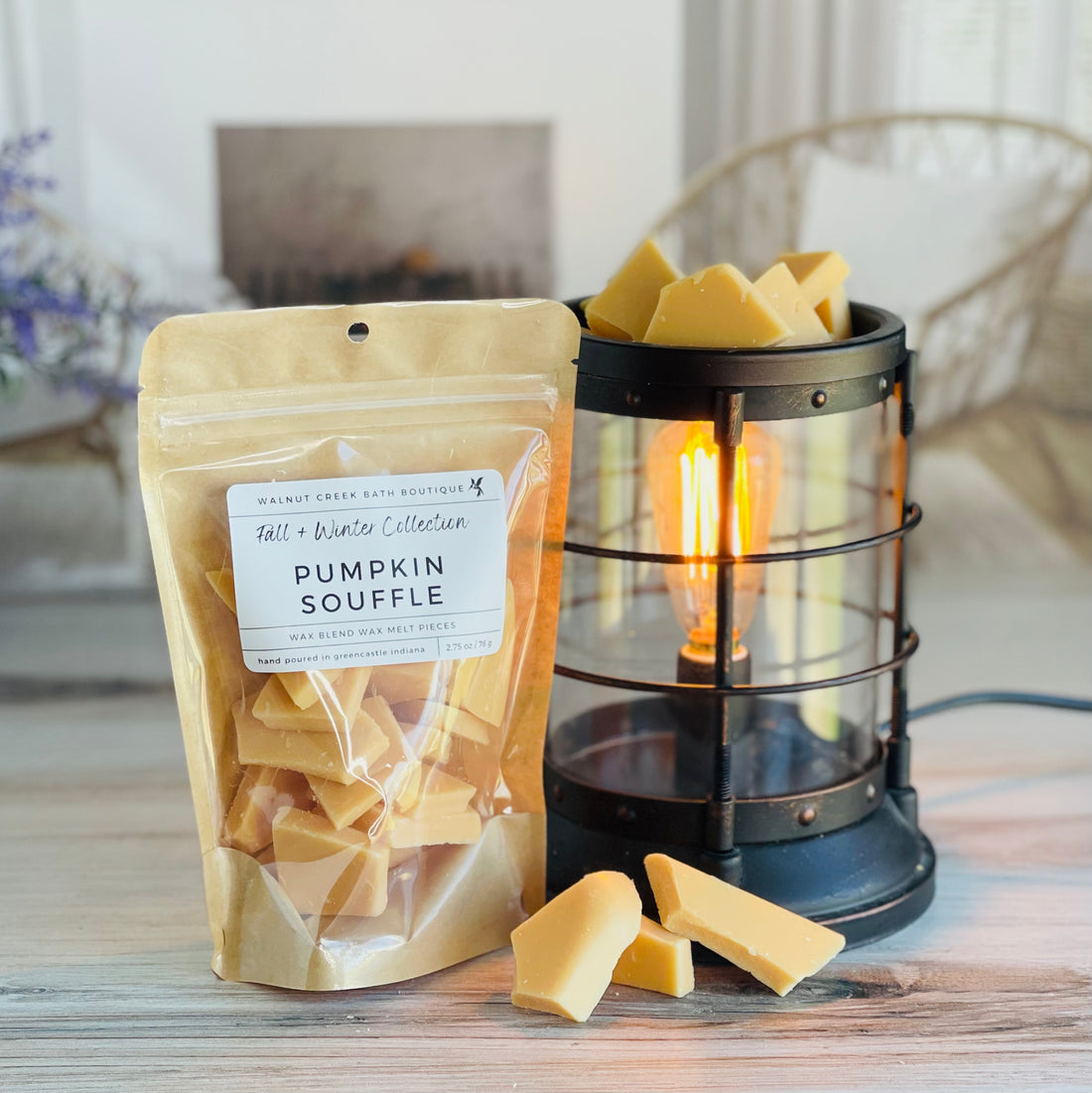 pumpkin souffle wax melt bag is full and standing next to a rustic farmhouse type burner that is on and filled with the orange brittle pieces of wax. there are also a few shards of the brittle on the table in front of both to show the size and color of the pieces