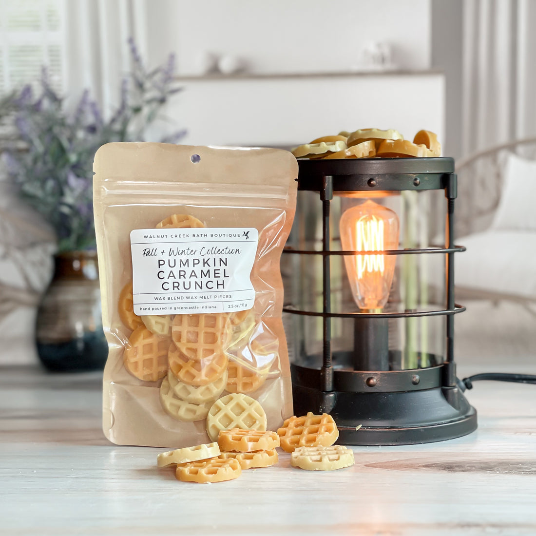 pumpkin caramel crunch wax melt bag next to lit burner. there are wax melts on the table scattered about to shoe the pieces - these are yellow and orange tiny waffle pieces