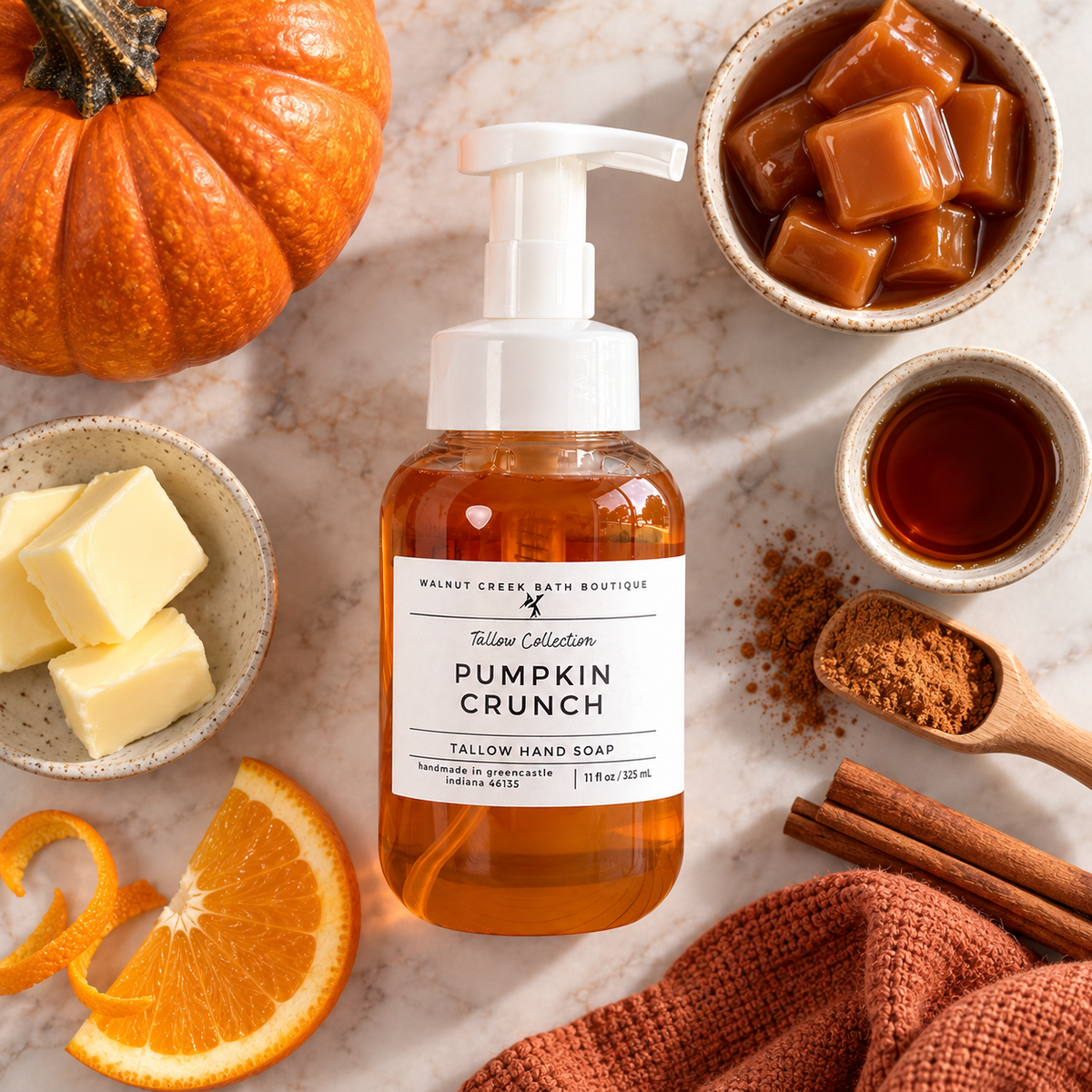 Walnut Creek Bath Boutique Pumpkin Crunch Tallow Hand Soap bottle, surrounded by seasonal ingredients including a whole pumpkin, butter, orange slices, cinnamon, and caramel pieces on a marble surface.