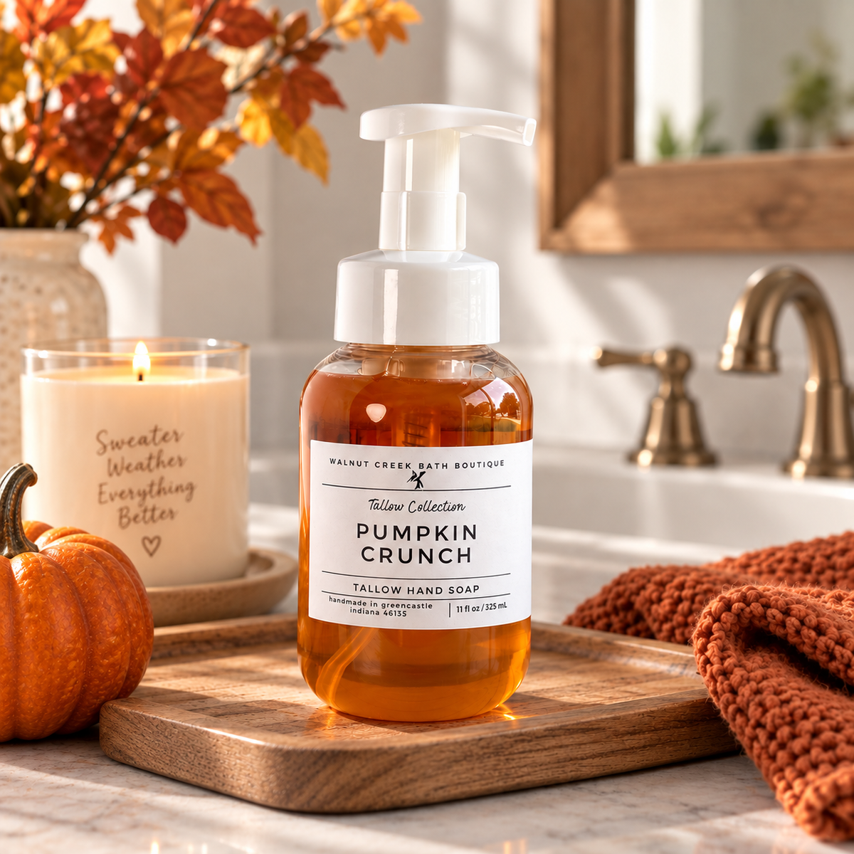 A bottle of Walnut Creek Bath Boutique Tallow Collection Pumpkin Crunch Tallow Hand Soap sits on a wooden cutting board in a warm, autumn-themed bathroom setting with a candle, small pumpkin, and knit blanket.