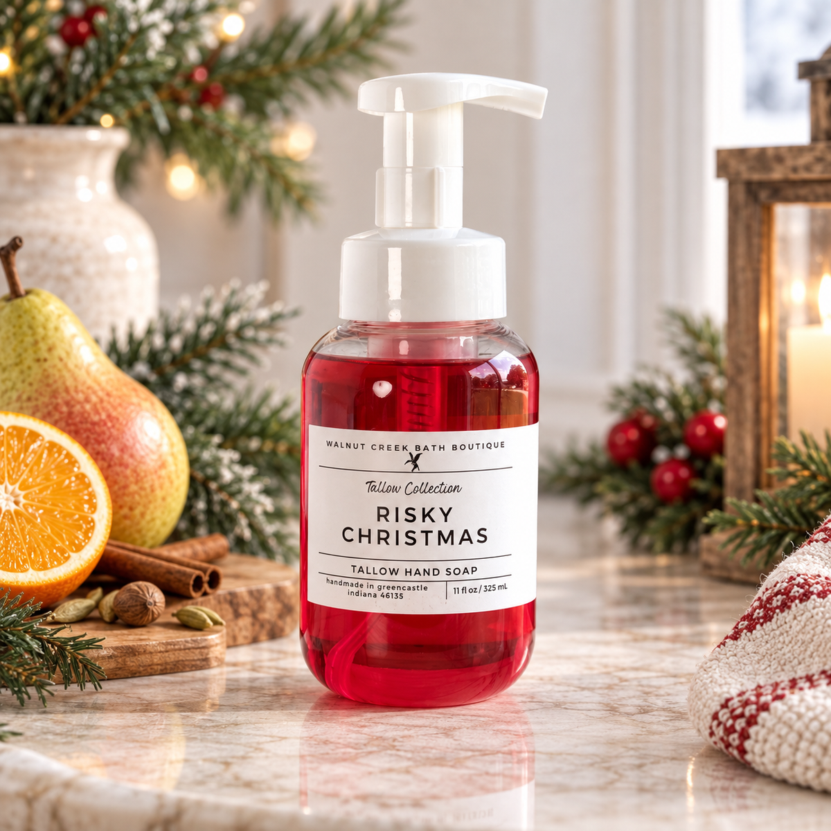 Walnut Creek Bath Boutique Risky Christmas Tallow Hand Soap in a clear pump bottle filled with red liquid, styled on a marble counter with holiday decorations including a pear, orange slice, cinnamon stick, pine branches, and a lit candle.