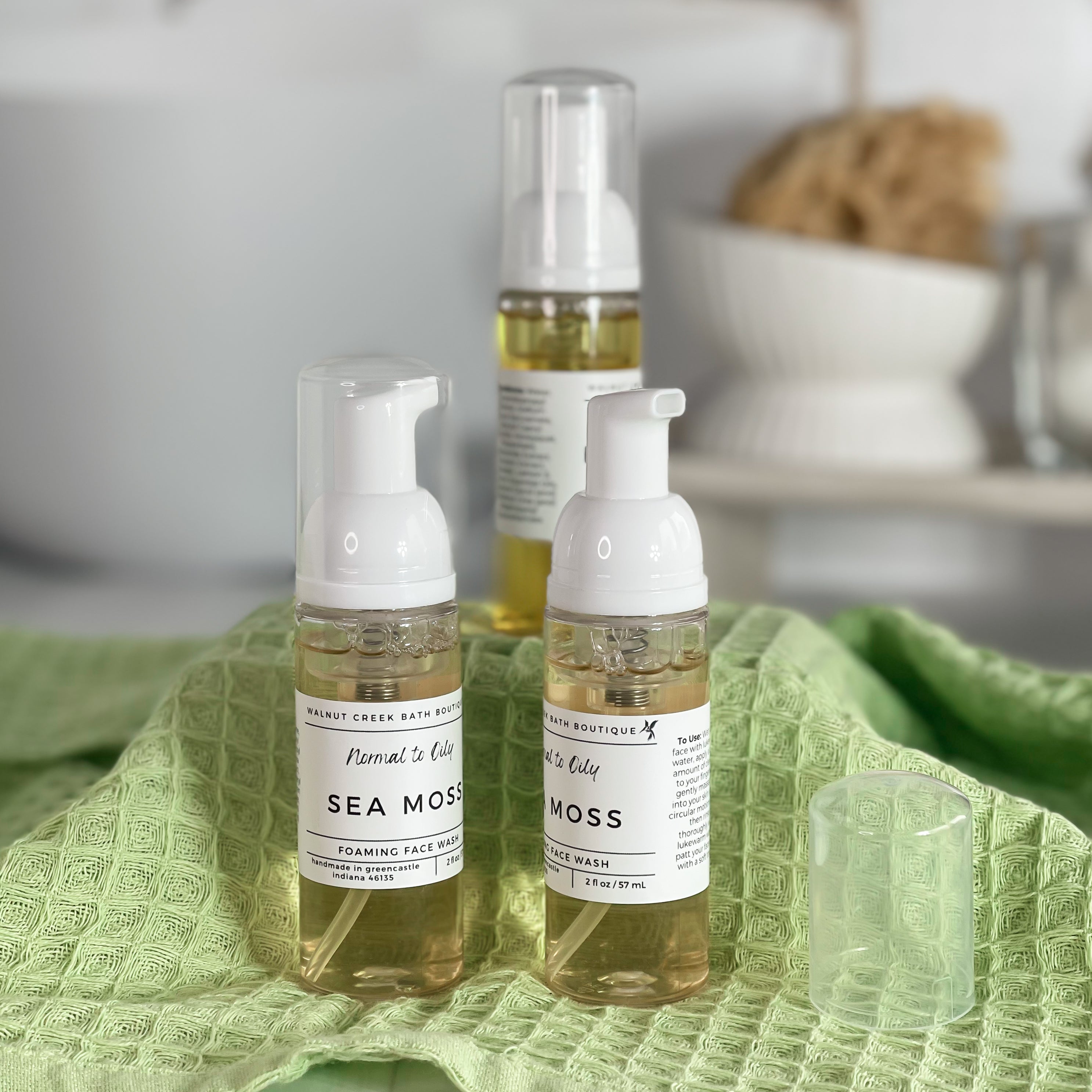 3 sea moss foaming face washes are shown on a green textured cloth covered riser. they are a light yellow color. in the background is a spa like bathroom