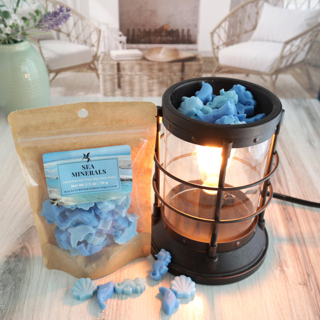 sea minerals wax melts are cute little pieces of wax. there are seashells, dolphins, seahorses and whales! some are lighter blue and some are darkers. the image has a bag of wax melts standing next to a lit burner full of these wax pieces while a sprinkling of them are also scattered on the table in front of the bag and burner.  this is all sitting on a washed out wooden base with a couple of chairs in the background next to a fireplace