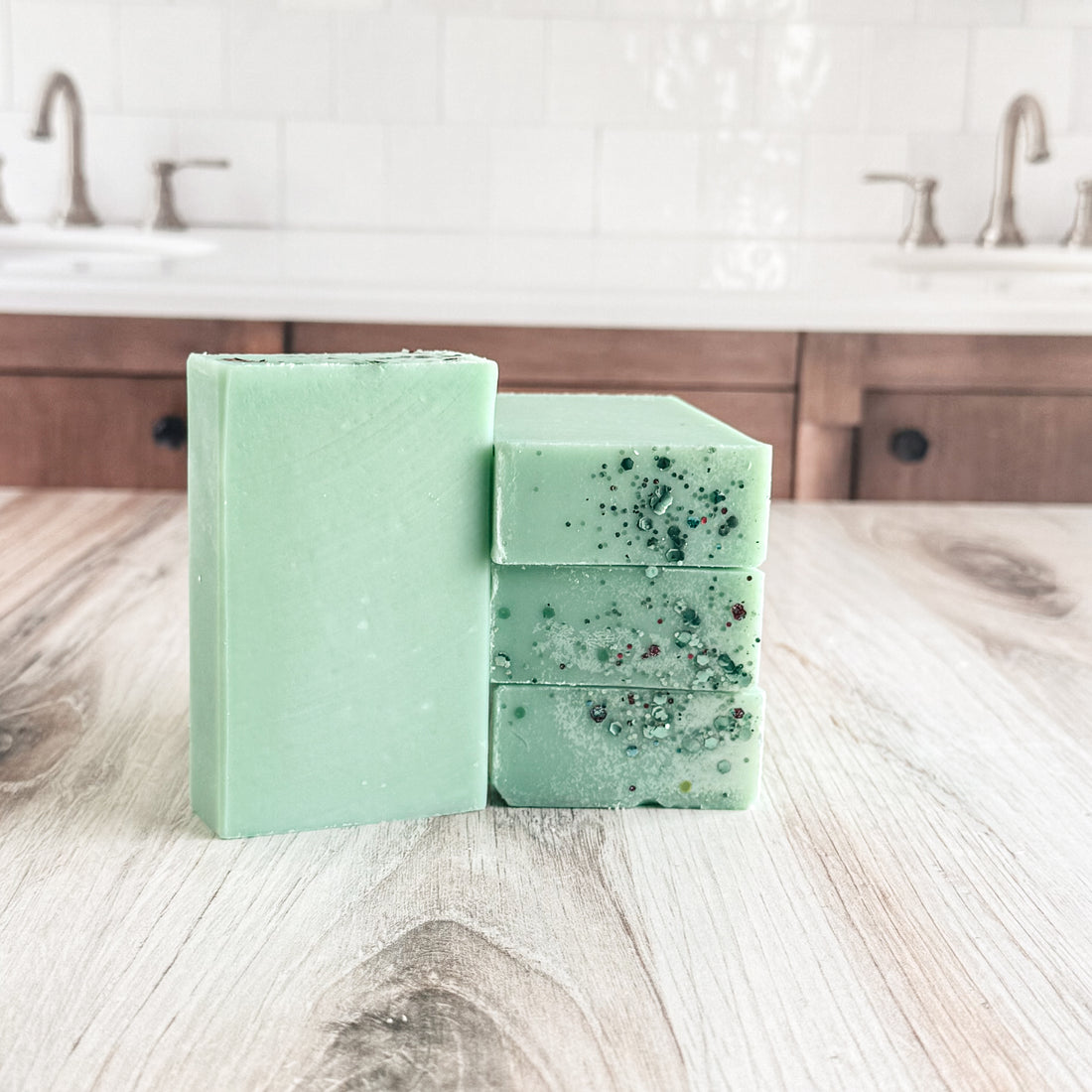 Three bars of handmade soap in a light mint green color, one standing upright and two stacked, with dark green and glittery speckles on the side bars, displayed on a light wood countertop with a blurred kitchen sink in the background.