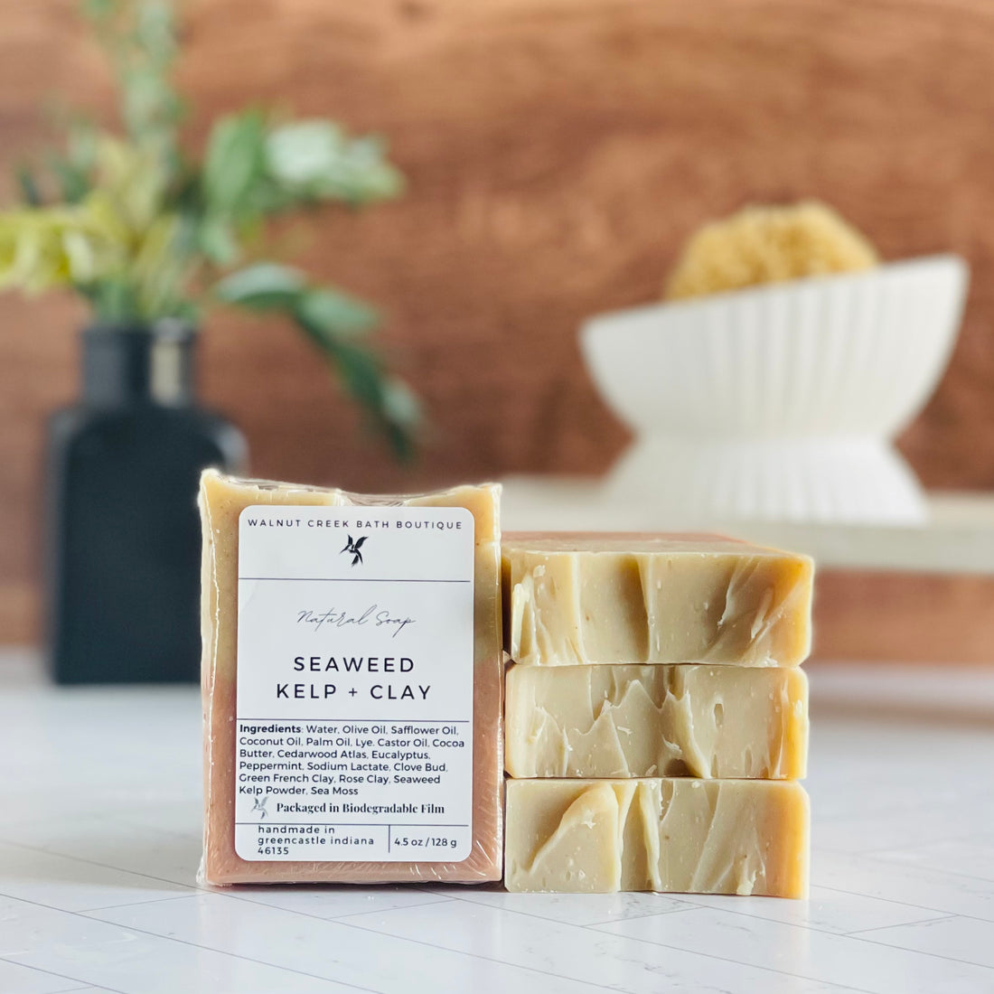 seaweed kelp and clay soaps are shown, one standing facing out to show the label. Next to this are three more soaps stacked on top to show the sculpted tops. in the background is a soap dish on a stand and a small vase with some greenery.