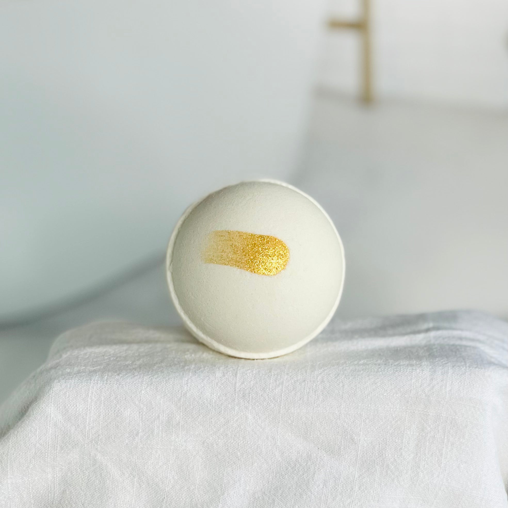 spa day bath bomb up close. this is a white bath bomb with a gold slash over it. it is sitting center stage on a white cloth over a riser