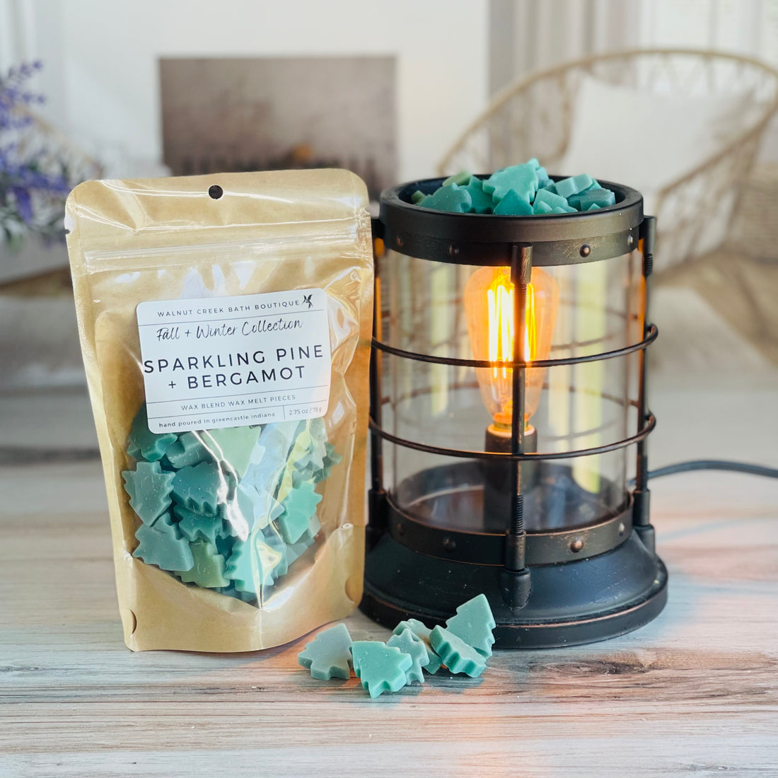 sparkling pine and bergamot wax melt bag is full and standing next to a rustic farmhouse type burner that is on and filled with the green tree pieces of wax. there are also a few trees on the table in front of both to show the size and color of the pieces