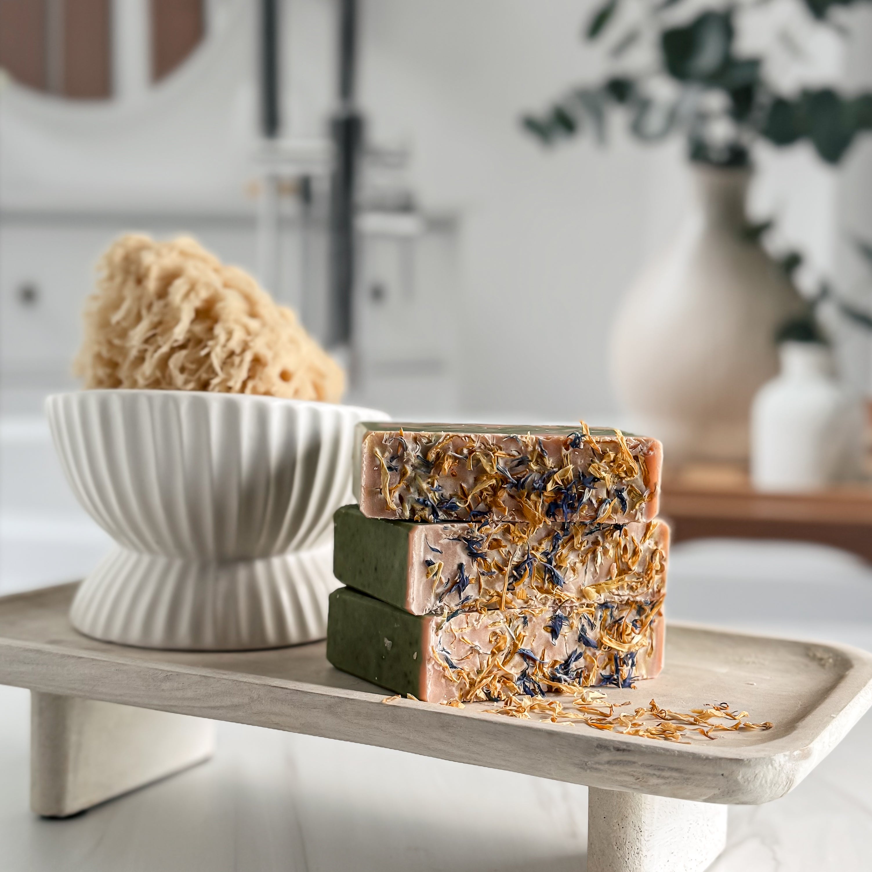 Three soap bars with natural ingredients on a wooden stand, with a bowl of sponges in the background.