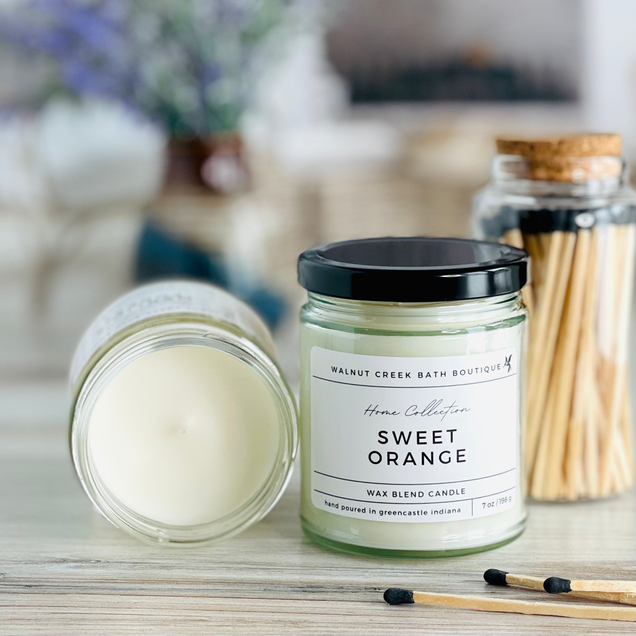 sweet orange candle with lid to the right of another candle with the top off to show the creamy wax inside. there are a few matches scattered in front and a pretty glass jar of matches in the background