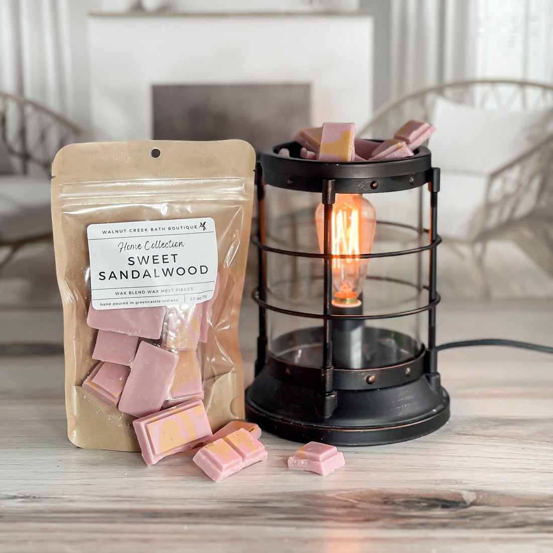 sweet sandalwood wax melt bag is full and standing next to a rustic farmhouse type burner that is on and filled with the pieces of wax in the shape of chocolate candy pieces - pink with a little yellow drizzle!. there are also some pieces on the table in front of both to show the size and color of the pieces