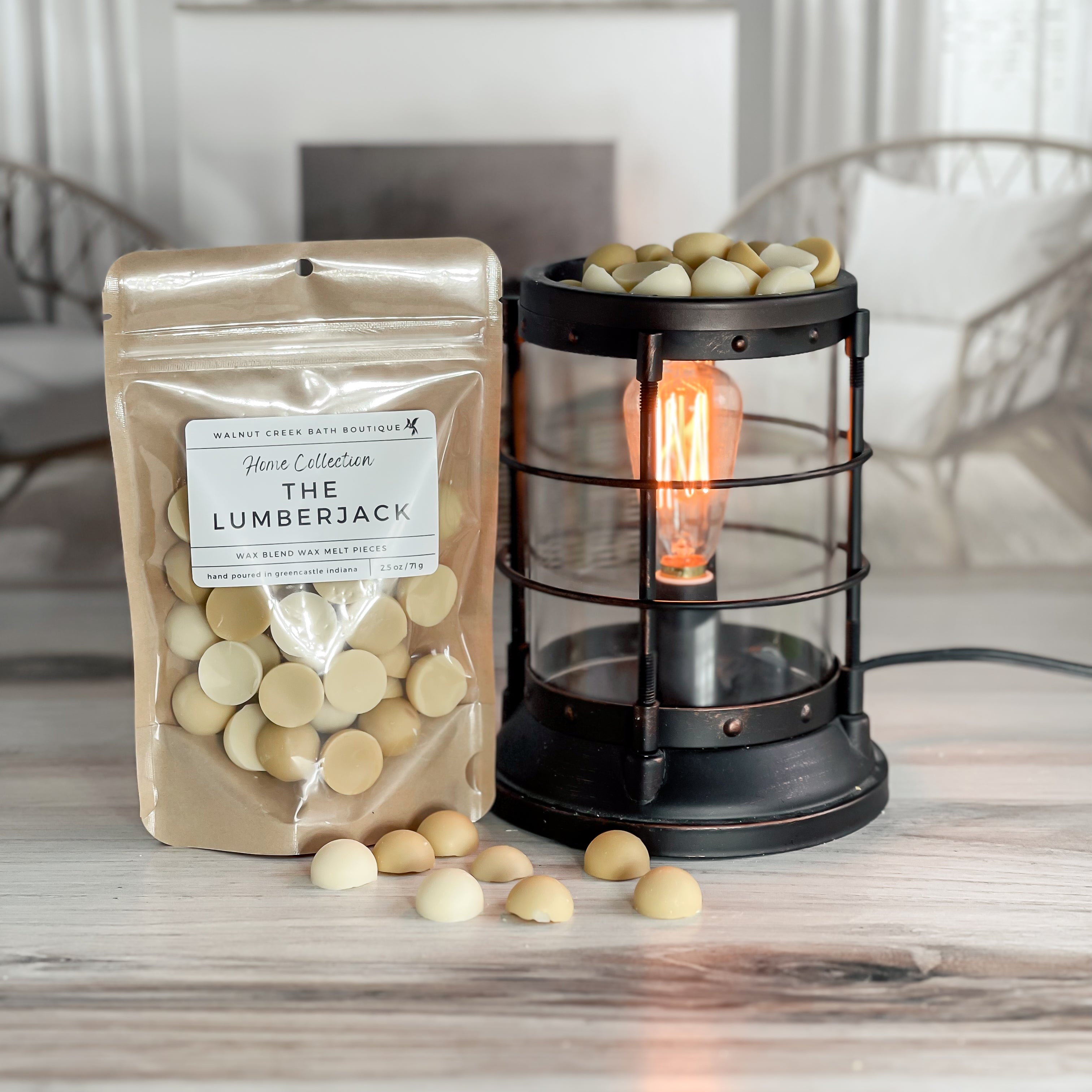 the lumberjack wax melt bag is full and standing next to a rustic farmhouse type burner that is on and filled with the light and dark tan half sphere pieces of wax. there are also a few rounds of the wax on the table in front of both to show the size and color of the pieces