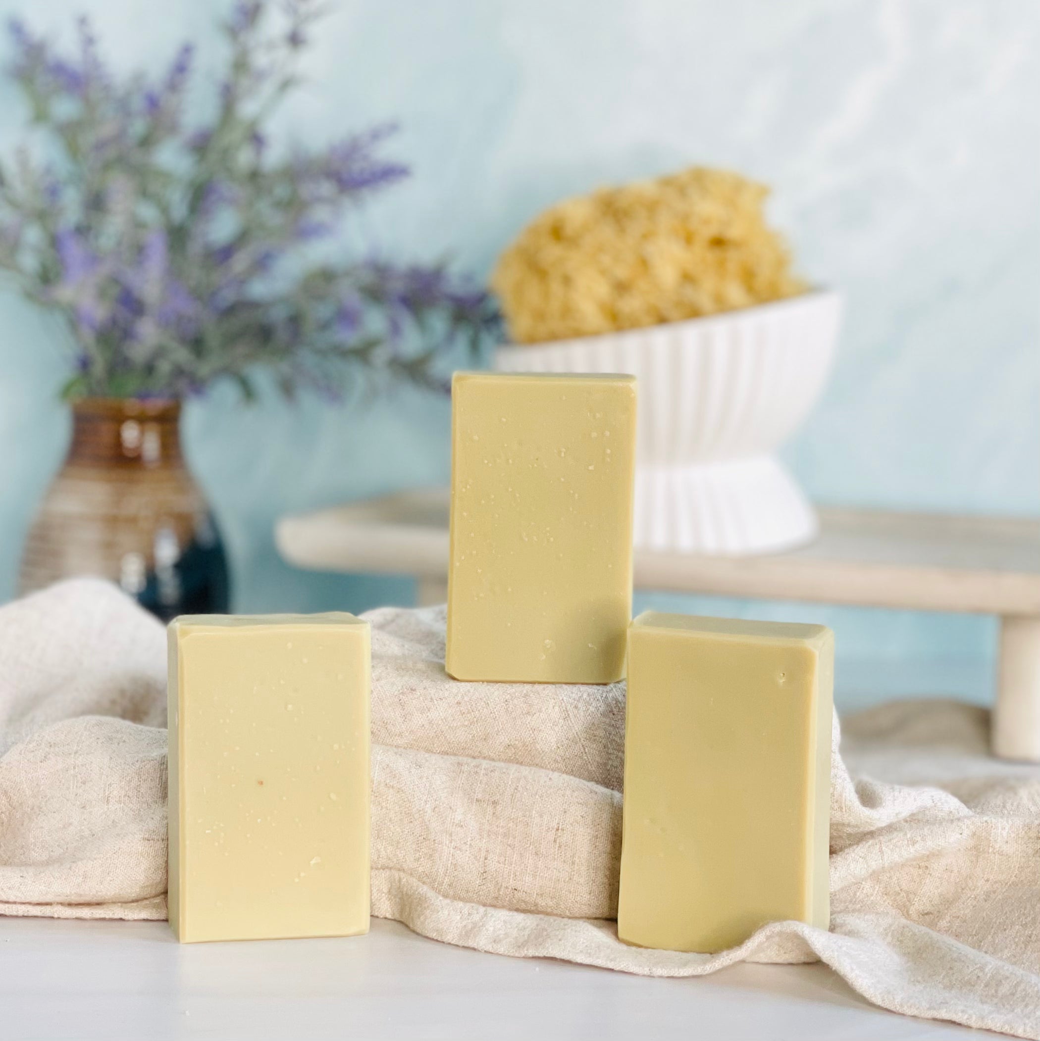 three aleppo style soaps are shown with one on a tan covered dias along with a loofah in a dish in the background