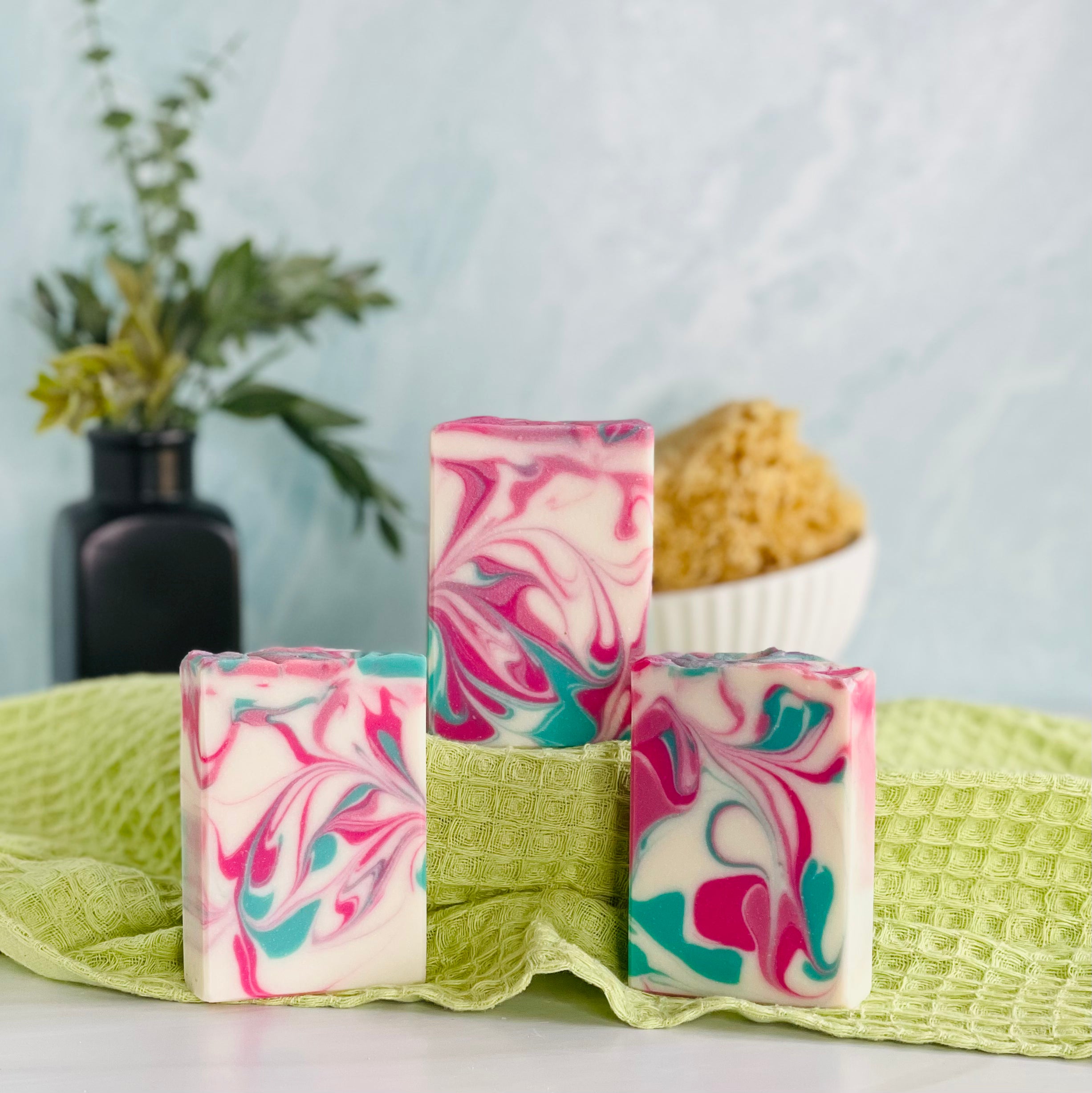 three blackberry magnolia soaps are shown with one on a green covered dias along with a loofah in a dish in the background