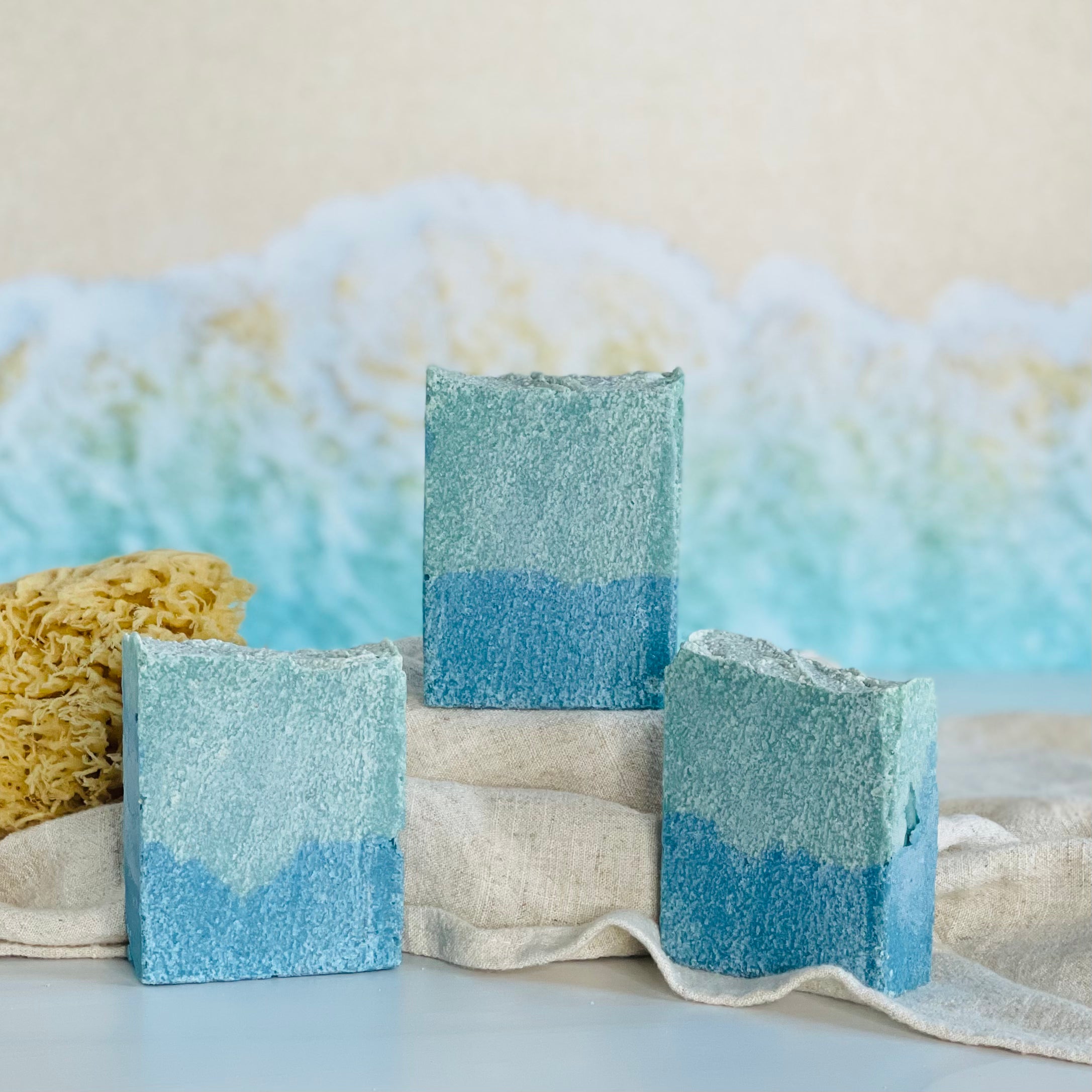 three flamingo beech soaps are shown with one on a tan covered dias along with a loofah in a dish in the background