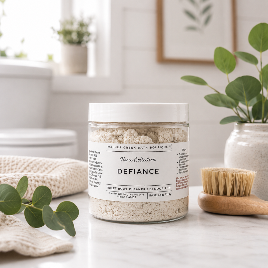 A clear plastic jar of walnut creek bath boutique Defiance toilet bowl cleaner and deodorizer sits on a marble countertop with a wooden brush and eucalyptus leaves in a bright bathroom setting.