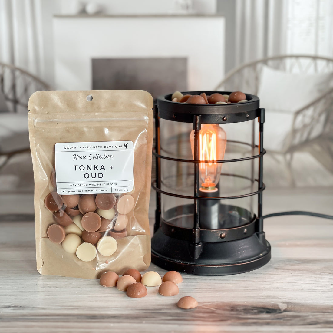 tonka and oud wax melt bag is full and standing next to a rustic farmhouse type burner that is on and filled with the light, medium and dark tan half sphere pieces of wax. there are also a few rounds of the wax on the table in front of both to show the size and color of the pieces