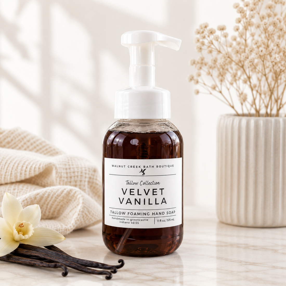 A bottle of Walnut Creek Bath Boutique Velvet Vanilla Fallow Foaming Hand Soap stands on a marble surface with a beige towel, vanilla beans, and dried flowers in the background.
