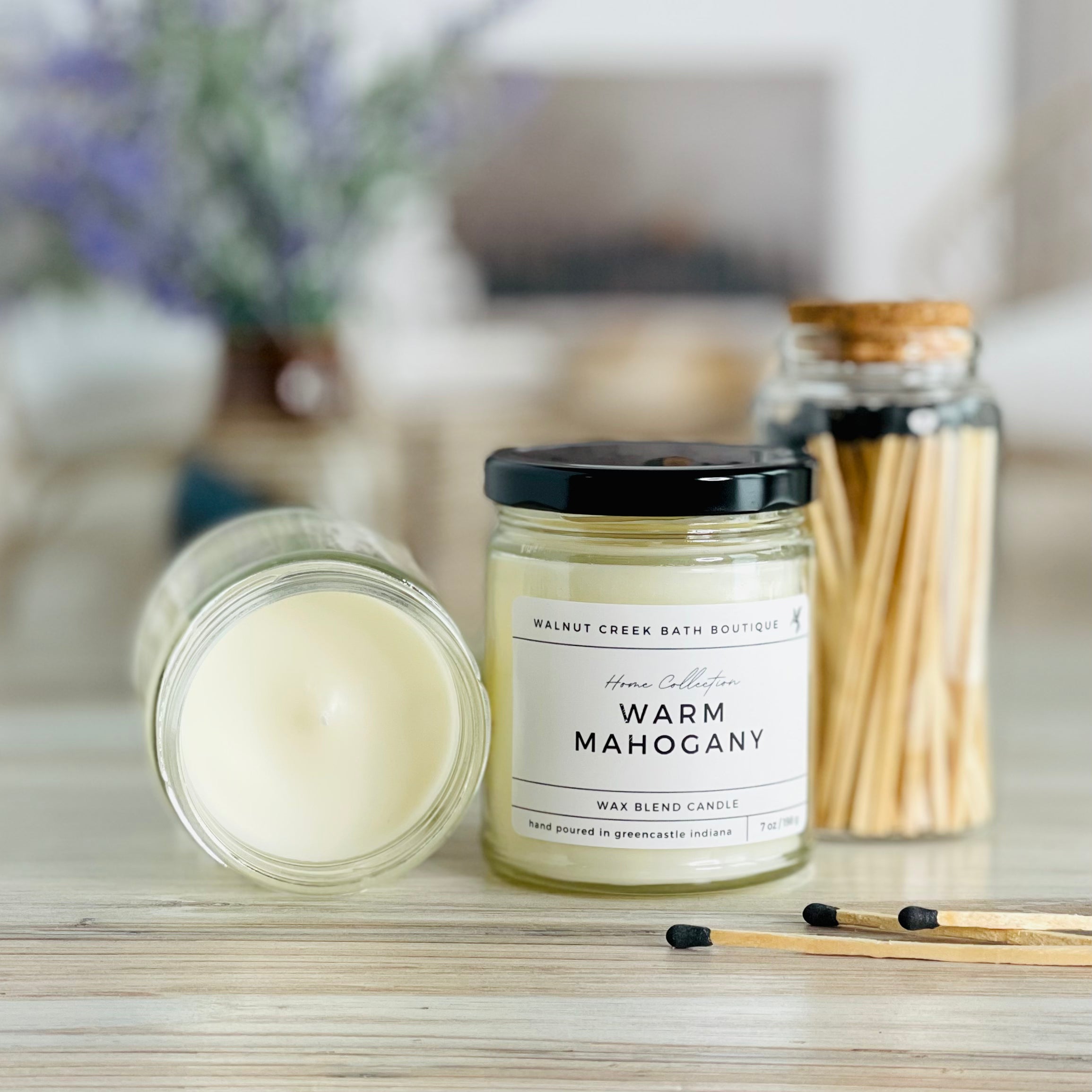 warm mahogany candle with lid to the right of another candle with the top off to show the creamy wax inside. there are a few matches scattered in front and a pretty glass jar of matches in the background