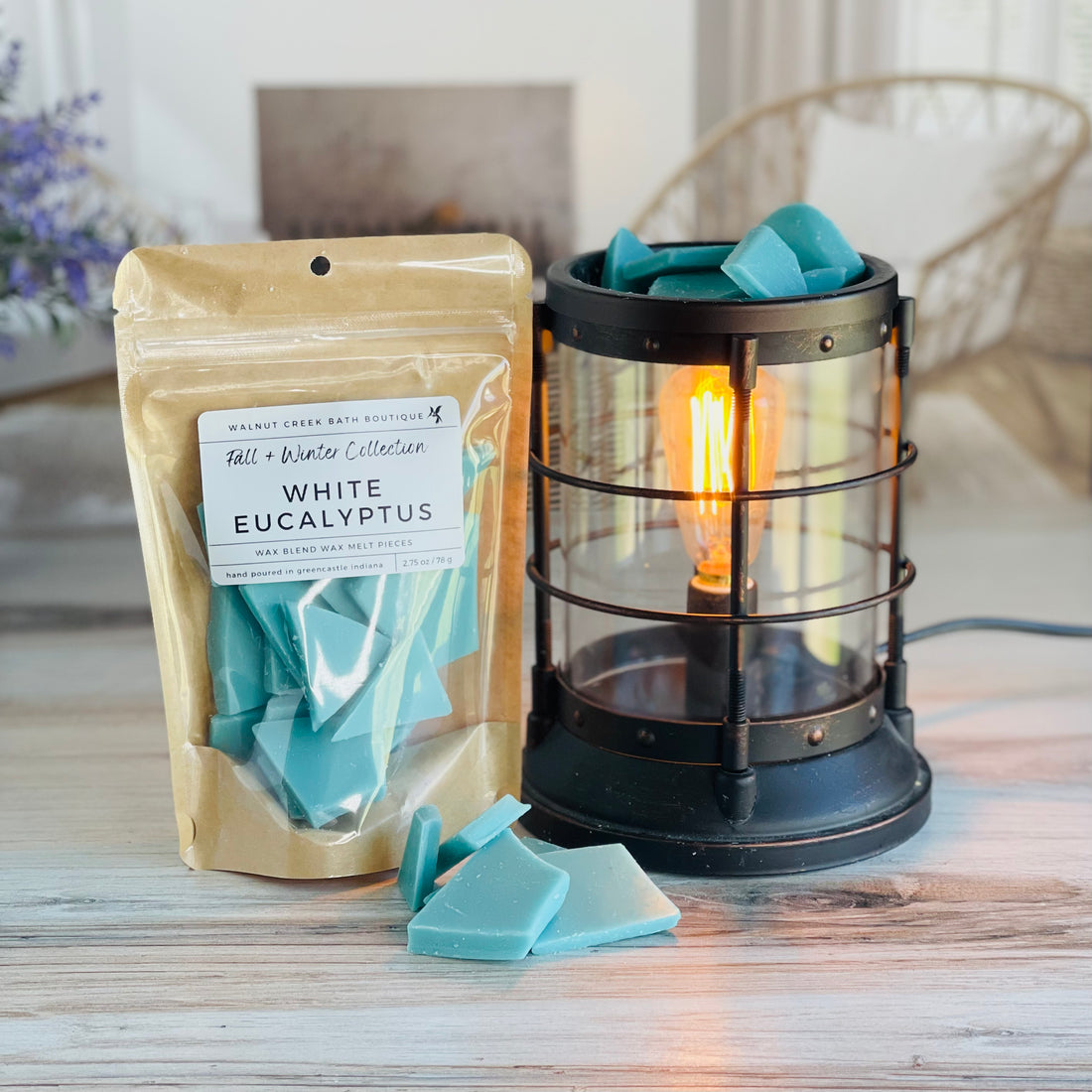white eucalyptus wax melt bag is full and standing next to a rustic farmhouse type burner that is on and filled with the mint green brittle pieces of wax. there are also a few shards of the brittle on the table in front of both to show the size and color of the pieces