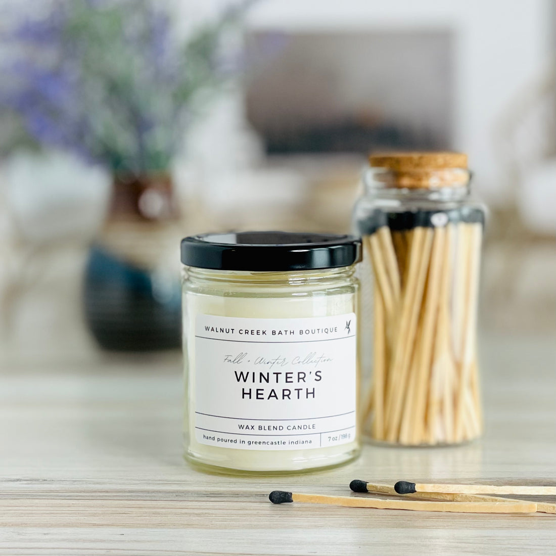 winters hearth candle with black lid. there are a few matches scattered in front and a pretty glass jar of matches in the background