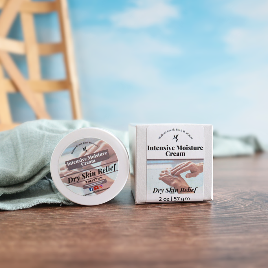 1 jar of intensive moisture cream is sitting on hits side next to another cream in its box to show packaging. They are sitting on a wooden board with some pink salts sprinkled around them. There is a sky background and a green towel throughout the scene
