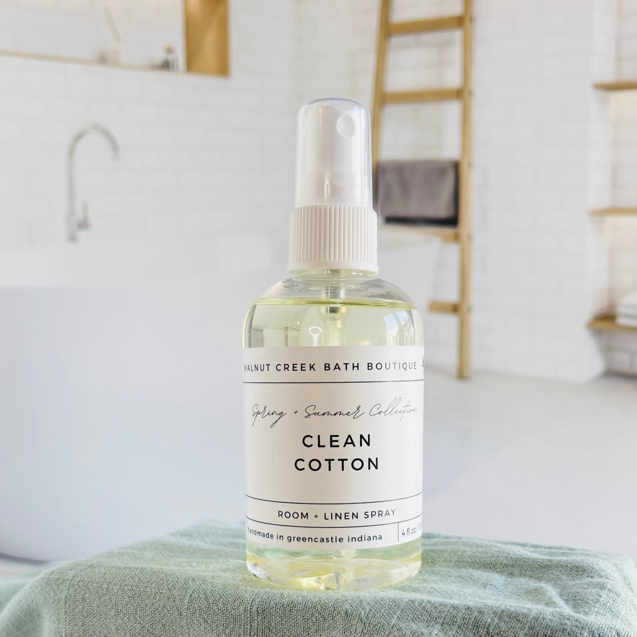 sea salt and lily room spray is shown . in the background is a second bottle on a tray next to a green vase with small flower buds. this is in front an image of a fireplace flanked by two chairs. there is some greenery hanging from the back right 