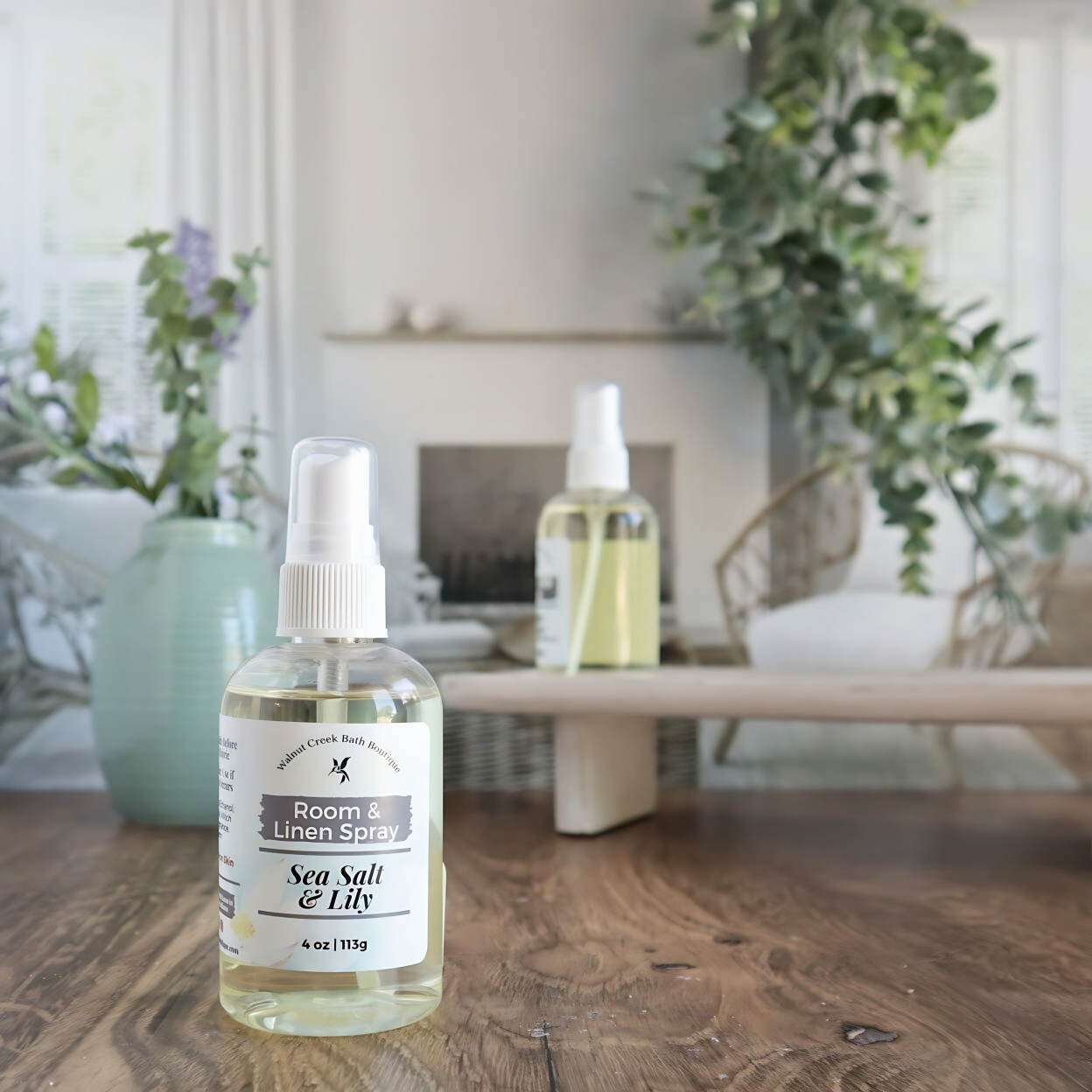 sea salt and lily room spray is shown . in the background is a second bottle on a tray next to a green vase with small flower buds. this is in front an image of a fireplace flanked by two chairs. there is some greenery hanging from the back right 