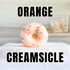 a single orange creamsicle bath bomb donut is shown. it is orange in color and is dipped in white bath glaze. then it has bath sprinkles scattered over the top. the words orange creamsicle is on the image.