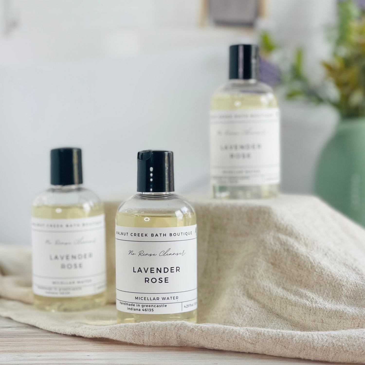 one lavender rose micellar water is lifted up on a tan box somewhat out of focus. there are 2 others in front of this box, with one being front and center and in sharp focus.  this is set in front of a spa like bathroom and in the background is a sage green vase holing some greenery 