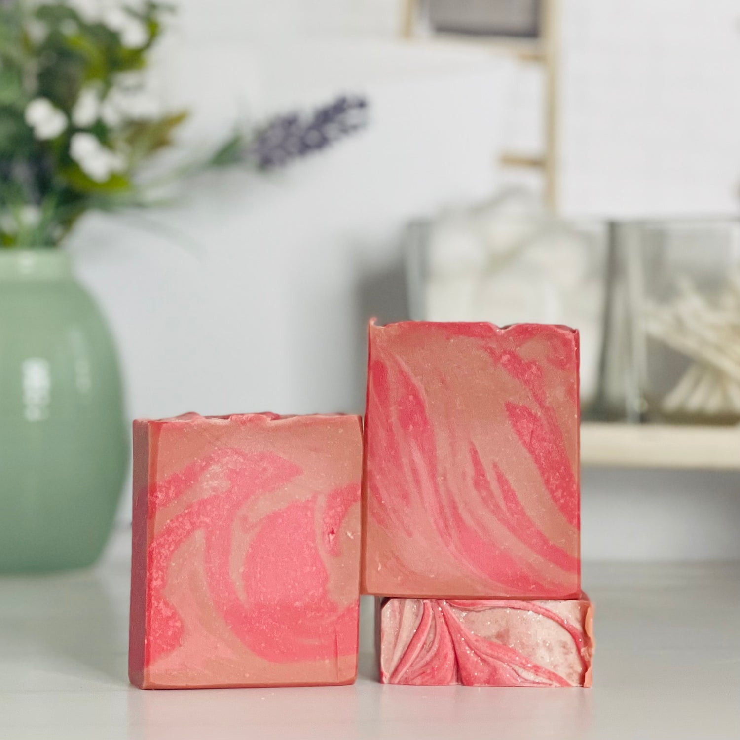 there are three vanilla kissed soaps, one laying flat to show the top and one standing tall on top of it. next to those is a third soap standing next to them showing the two pinks and the swirls. in the background is a sage green vase with some small buds.