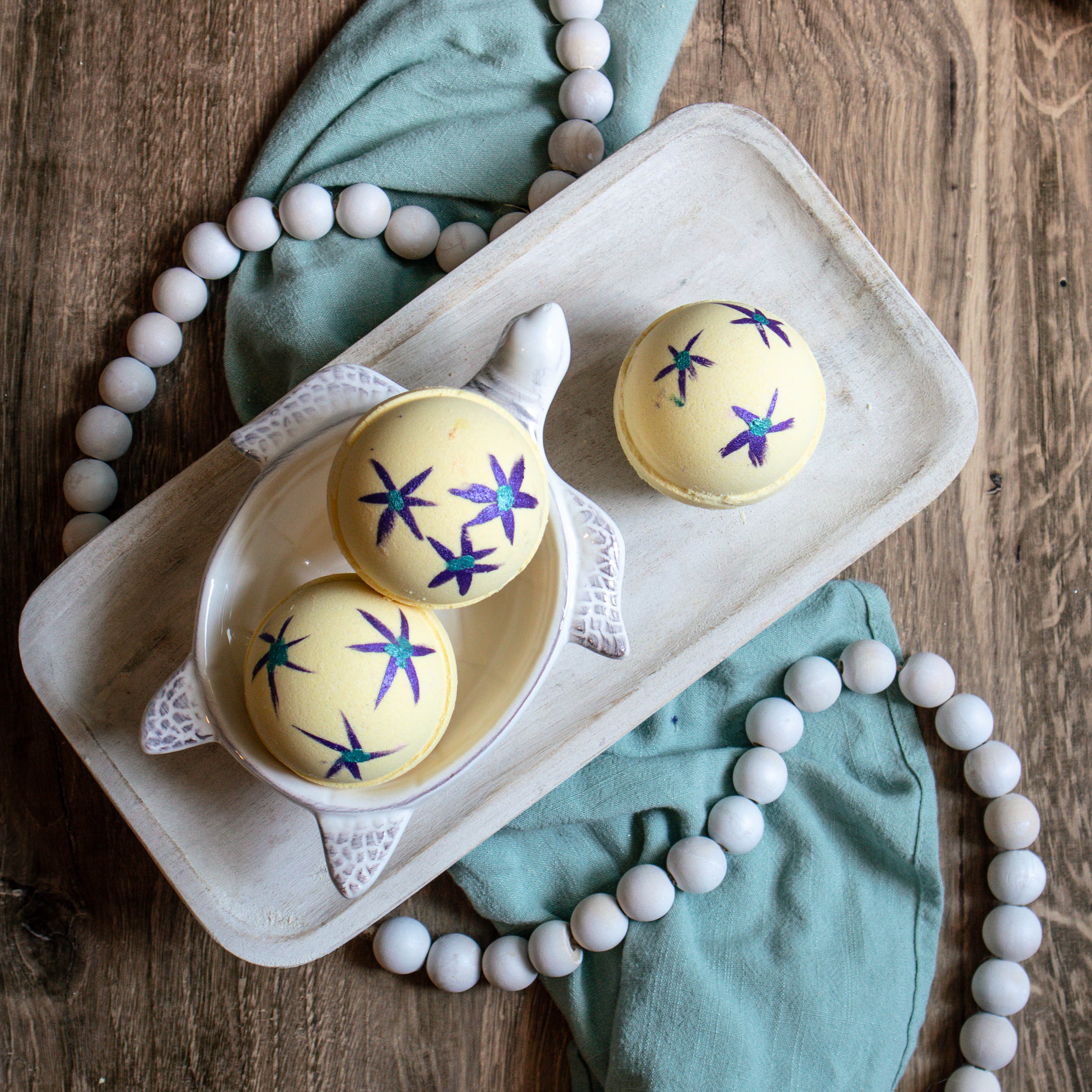 there are 3 yellow bath bombs, 2 are in a turtle shaped soap dish and one is off to the side on its own. these bath bombs have purple flowers with green centers painted on them. there is a green towel running along beneath the tray and some white beads strung along beneath the tray as well