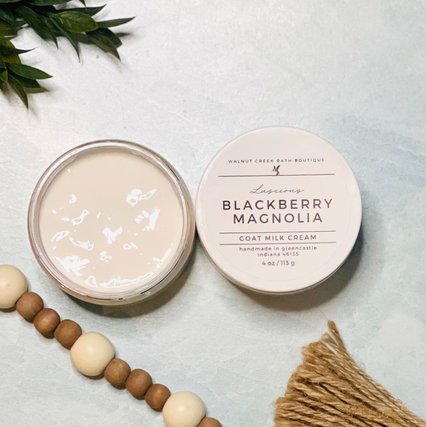 4 oz blackberry magnolia goat milk cream open with the lid next to it. the texture is so creamy and luscious looking. there is some greenery on the top left and some wooden beads running along the edge with the tassel peaking just in view on the bottom right.