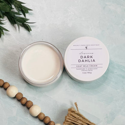 4 oz dark dahlia goat milk cream open with the lid next to it. the texture is so creamy and luscious looking. there is some greenery on the top left and some wooden beads running along the edge with the tassel peaking just in view on the bottom right.