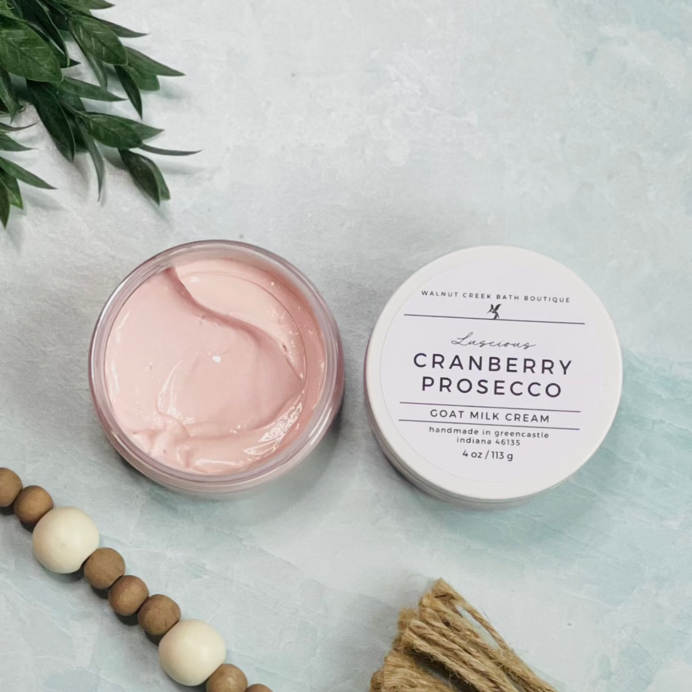 7 oz cranberry prosecco goat milk cream open with the lid next to it. the texture is so creamy and luscious looking. there is some greenery on the top left and some wooden beads running along the edge with the tassel peaking just in view on the bottom right.