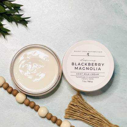 7 oz blackberry magnolia goat milk cream open with the lid next to it. the texture is so creamy and luscious looking. there is some greenery on the top left and some wooden beads running along the edge with the tassel peaking just in view on the bottom right.