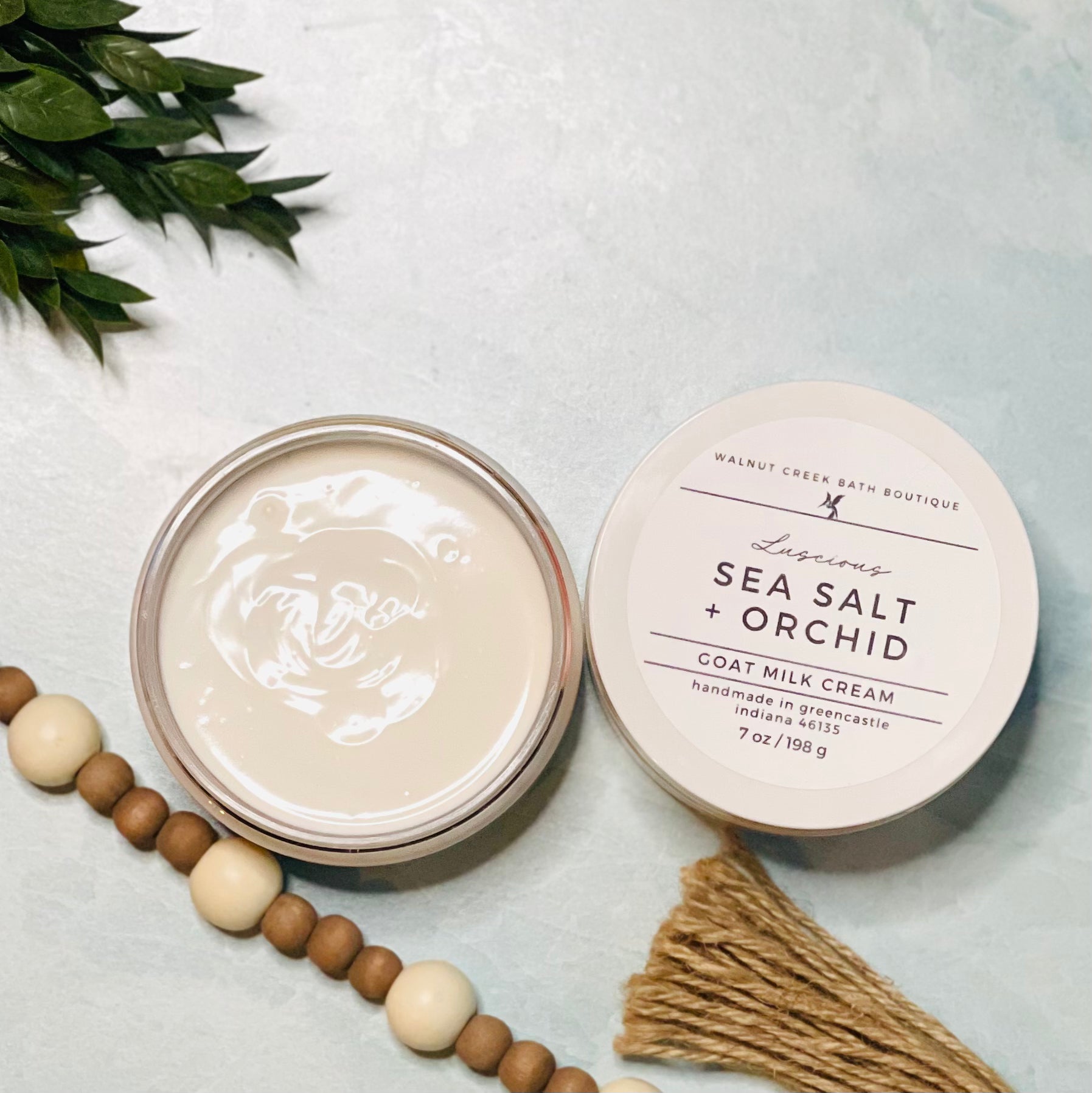 7 oz sea salt and orchid goat milk cream open with the lid next to it. the texture is so creamy and luscious looking. there is some greenery on the top left and some wooden beads running along the edge with the tassel peaking just in view on the bottom right.