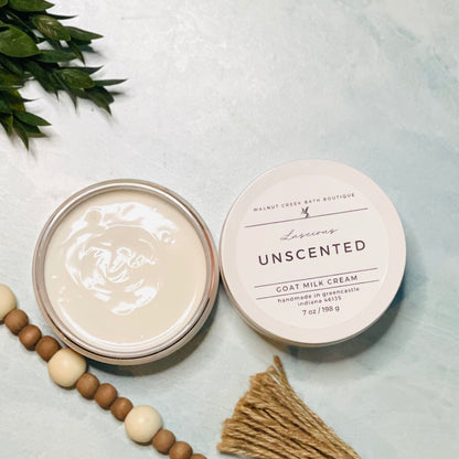 7 oz unscented goat milk cream open with the lid next to it. the texture is so creamy and luscious looking. there is some greenery on the top left and some wooden beads running along the edge with the tassel peaking just in view on the bottom right.