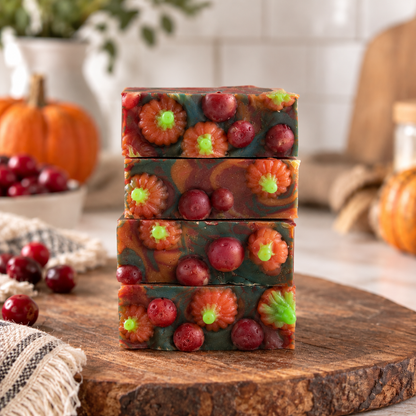 Four stacked rectangular bars of handmade soap with a dark green base, orange and red marbled swirls, and embedded cranberries and small orange pumpkins, displayed on a wooden board with a fall-themed background.