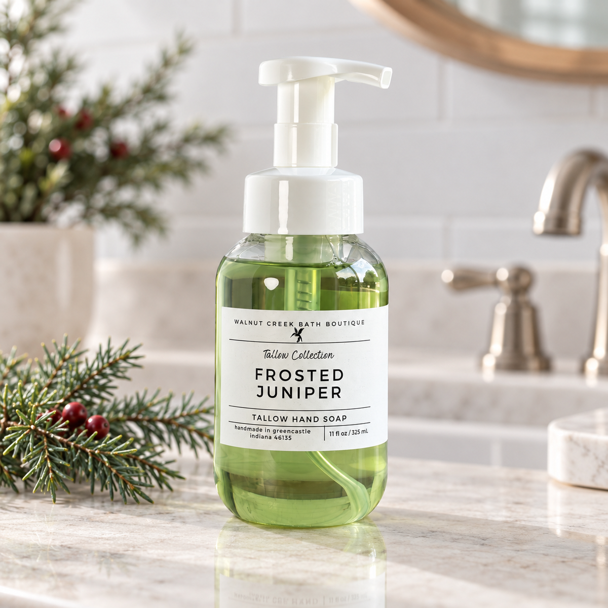 A bottle of Walnut Creek Bath Boutique Frosted Juniper yellow hand soap stands on a marble bathroom counter with pine branches nearby and a blurred sink and mirror in the background.