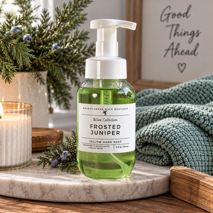 A bottle of Walnut Creek Bath Boutique Frosted Juniper hand soap sits on a marble tray in a cozy, styled setting with a lit candle, pine branches, a knit blanket, and a sign that reads &