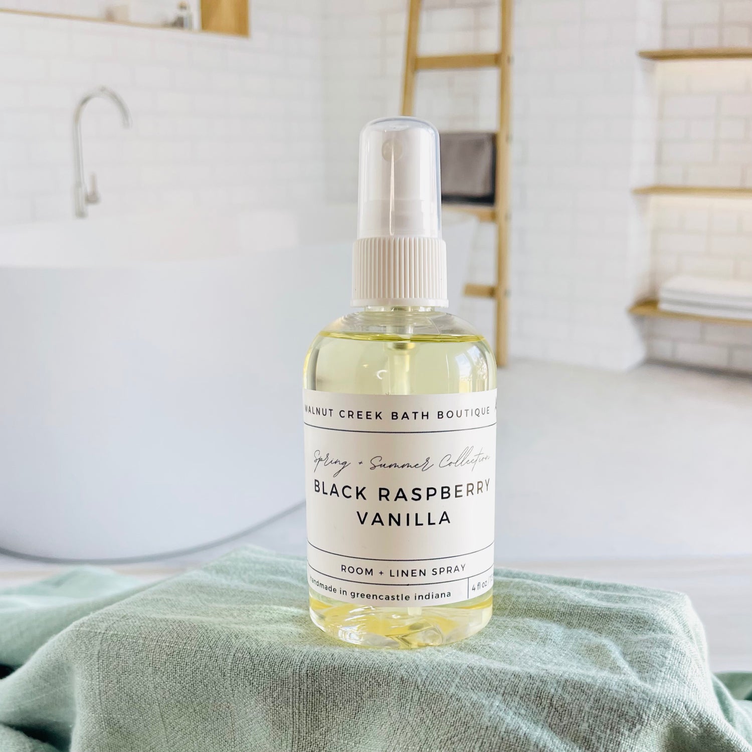 black raspberry vanilla room and linen spray is in a clear bottle with a white spray nozzle. it is sitting on a raised platform with a green towel unter it and in the background is a spa like bathtub