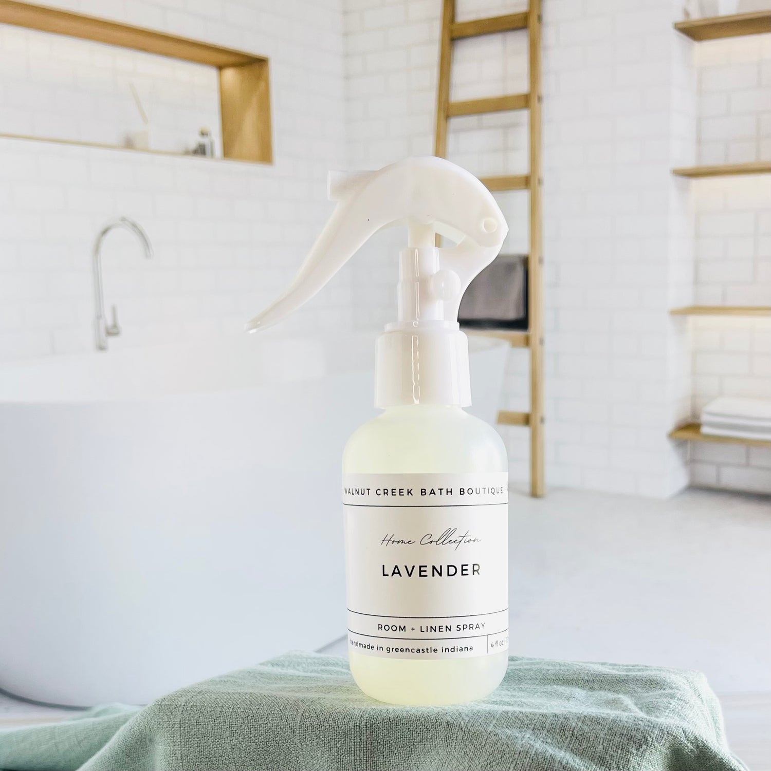 lavender room and linen spray is in a opaque bottle with a larger spray nozzle for ease of use. it is sitting on a raised platform with a green towel unter it and in the background is a spa like bathtub