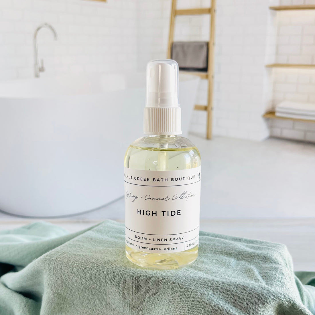 high tide room and linen spray is in a clear bottle with a white spray nozzle. it is sitting on a raised platform with a green towel unter it and in the background is a spa like bathtub